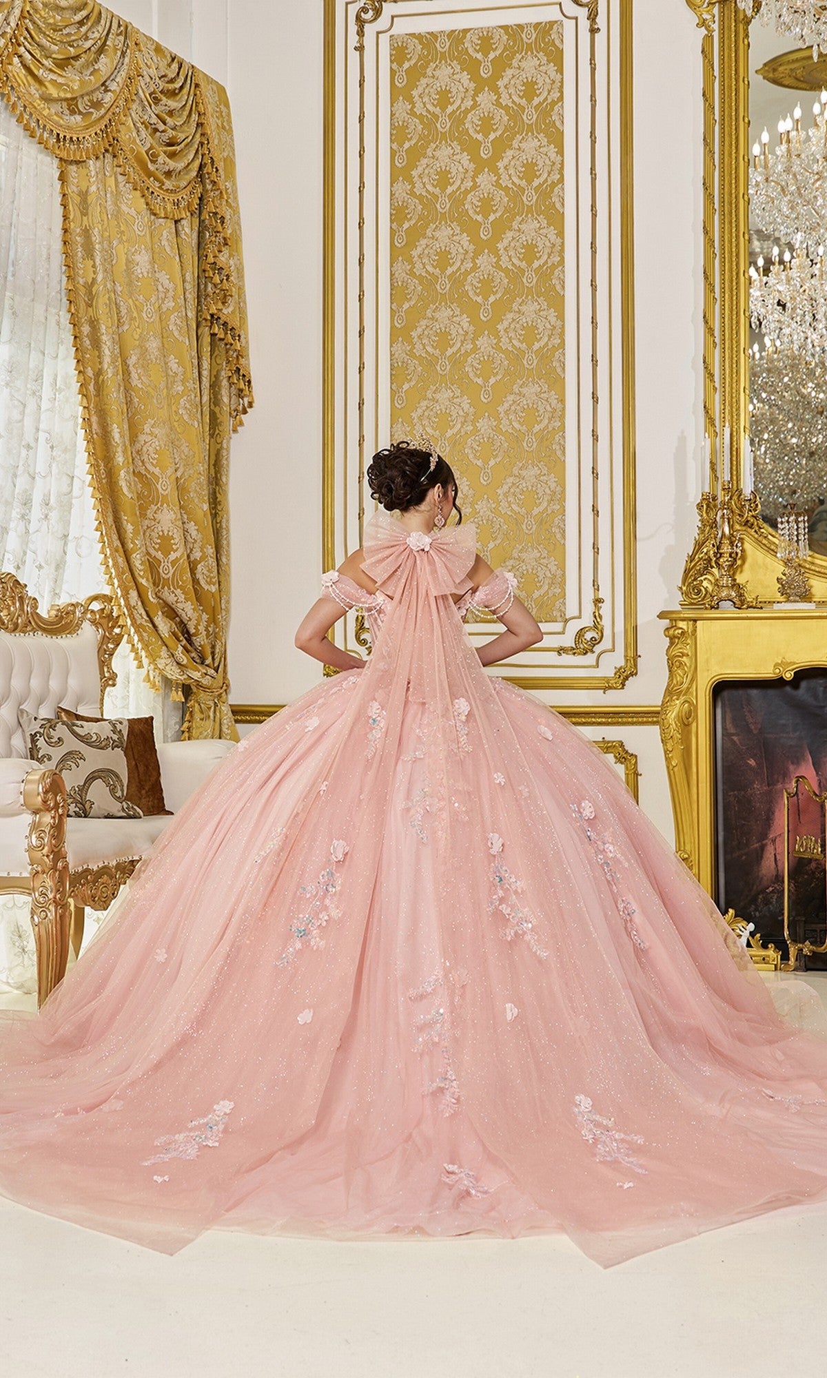 A person in the Quinceanera Dress 5096 By Dancing Queen, featuring blush pink glitter tulle and floral embellishments, stands before an ornate gold-trimmed wall in a lavishly decorated room.